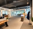 Modern office space with a reception area, desks, chairs, and a coat rack. The room features a sleek design with glass partitions and pendant lighting.