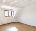 Empty attic room with wooden floors, white walls, exposed wooden beams, and a single window with wooden frame.