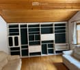 Living room with light wooden flooring, white built-in bookshelf with multiple compartments and cabinets, two beige sofas, and a window with white curtains.