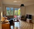 Bright living room with wooden parquet flooring, large windows and sliding doors leading to a balcony, a sofa, armchair, coffee table, TV, and table lamp.