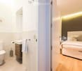Modern bedroom with a white bed, nightstand, and lamp. Adjacent bathroom features a toilet, sink, and cabinet.