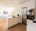 Modern kitchen with white cabinets, stainless steel appliances, and a central island. The space features a sink, microwave, oven, and a cup on the counter. A picture frame is visible on the wall, and the floor is wooden.