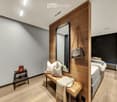 Modern bedroom with wooden flooring, a large mirror, a bench with pillows and a blanket, a small stool with shoes, and a bed with pillows and a blanket.
