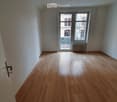 Empty room with wooden flooring, white walls, and a large window with a view of a building and balcony.