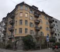 Multi-story residential building with yellow facade, multiple balconies, and grey shutters. The building has a rounded corner and is surrounded by greenery and parked motorcycles.