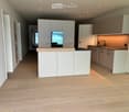 Modern kitchen with white cabinets, wooden countertops, and a central island. The floor is made of light wooden planks, and the space is well-lit with recessed lighting.