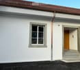 Two-story building with white exterior, red-tiled roof, and a small balcony on the roof. Features a window with a grey frame and a wooden door with a small step leading up to it.