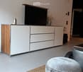 Modern living room with a grey ottoman, a sleek white and wood TV stand, a flat-screen TV, and decorative vases on the stand.