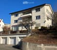 A multi-story white house with balconies, large windows, and a garage. The house is situated on a hillside with a stone retaining wall and a driveway leading to the garage.