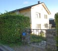 A two-story house with white walls and multiple windows, partially obscured by greenery and a stone gate with the number 6.