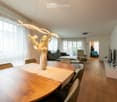 large windows, white walls, modern chandelier, wooden floor, modern furniture, dining table, chairs, living room, couch, center table, TV, pictures