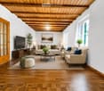 Modern living room with wooden floors, exposed wooden ceiling beams, large windows, and a mix of neutral and blue tones. Features a sofa, coffee table, ottoman, armchair, side tables, lamps, and a television.