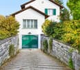 A two-story white house with green shutters and a green front door, surrounded by lush greenery and a stone pathway leading to the entrance.