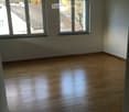 Empty room with light wooden flooring and large windows offering views of neighboring rooftops.