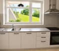 Modern kitchen with white cabinets, stainless steel sink, oven, induction cooktop, and range hood. Large windows offer a view of a green lawn and garden.