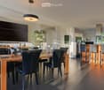 Modern dining room with a wooden dining table, black leather chairs, and grey bar stools. A large flat-screen TV is mounted on the wall, and a hanging pendant light illuminates the space. The room features a sleek, contemporary design with a mix of dark a