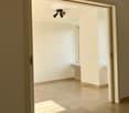 Empty room with beige tiled floor, white walls, and a small window letting in natural light. The room has a modern ceiling light fixture and a radiator near the window.