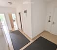 Bright hallway with white walls, tiled floor, and two black doormats. Doors lead to adjacent rooms, and a small mirror is mounted on the wall.