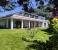A well-maintained lawn with a modern two-story house featuring a red-tiled roof and white columns, surrounded by lush greenery and flowering trees.