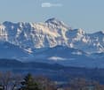 Panoramic view of snow-capped mountains and a valley with scattered trees and a body of water in the foreground.