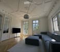 Elegant living room with ornate ceiling, large windows, grey sectional sofa, ottomans, flat-screen TV, and a modern floor lamp.