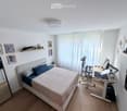 A modern, well-lit bedroom with a bed, desk, and shelves. The room features a large window with white curtains, a sleek desk setup with a computer, and decorative shelves with framed pictures and small plants.