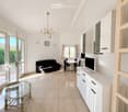 Bright living room with white walls, light wooden flooring, and large windows. Features a black sofa, glass coffee table, dining table with chairs, TV on a white cabinet, and a chandelier.