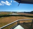 Balcony with metal railing overlooking a grassy field, road, and distant countryside with trees and mountains under a clear blue sky.