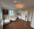 Modern, compact living space with a light-colored sectional sofa, a small round coffee table, a fitted kitchen area with cabinets and a fridge, and a large mirror on the wall. The room features wooden flooring and large windows allowing natural light.