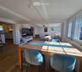 Bright living and dining area with wooden flooring, large windows, a sofa, a dining table with chairs, and a television on a stand.