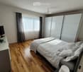 Modern bedroom with wooden flooring, large bed with white bedding, wardrobe, and large window with curtains.