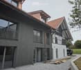 Modern two-story house with dark wooden siding, large windows, and a balcony on the upper level. The house features a red-tiled roof and a contemporary design with a mix of dark and light elements.