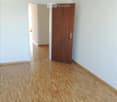 Empty room with light-colored wooden flooring and white walls. A wooden door is visible on the right side, and an open doorway leads to another room in the background.