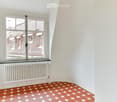 Empty room with white walls, hexagonal tile floor in shades of pink and red, large windows with view of rooftops, radiator below windows.