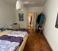 Bedroom with wooden flooring, a bed with a purple and white bedspread, a nightstand, a picture frame on the wall, and an open doorway leading to another room with a chair and table visible.