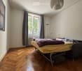 Bright bedroom with wooden herringbone flooring, large window with grey curtains, bed with yellow and purple bedding, radiator, picture frame, and a hanging lamp.