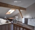 Loft-style room with exposed wooden beams, skylight windows, a sofa, a dining table with chairs, and a small kitchen area with cabinets and a dishwasher.