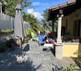 A well-maintained patio area with stone tiles, outdoor furniture including chairs and a table, potted plants, and an umbrella. The area is surrounded by greenery and has a view of mountains in the background.