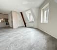 An unfurnished room with grey tiled flooring, white walls, and large windows. The room features a slanted ceiling and a small kitchen area with cabinets and appliances visible in the background.
