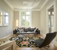 Spacious living room with large windows, hardwood floors, black leather sofa, patterned area rug, and decorative wall art