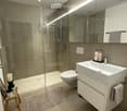 Modern bathroom with a glass-enclosed shower, white sink, toilet, and built-in vanity with mirrored cabinetry. Includes a small wooden stool with a candle and a beige bath mat.