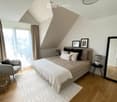 Modern bedroom with a large bed, grey armchair, side table, mirror, and decorative picture frames. The room features wooden flooring, a white rug, and large windows with city views.