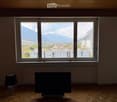 Empty room with wooden flooring, large windows offering a scenic mountain view, a table lamp, and a television on a stand.