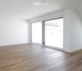 Empty attic room with wooden flooring, white walls, and large sliding glass doors leading to a view of the outside.