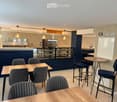 Modern pizzeria interior with wooden tables, upholstered chairs, bar stools, and a visible kitchen area with an oven and a sink.