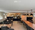 Modern restaurant interior with wooden tables, grey upholstered chairs, pendant lights, and a visible kitchen area with an oven.