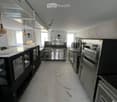 Commercial kitchen with stainless steel appliances, including a refrigerator, oven, and sink, along with a counter and pantry. The space features pendant lighting and large windows.