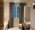Modern bathroom with white tiles, a bathtub, toilet, sink, and a wooden vanity. Features include a large window with blinds, a decorative vase with flowers, and a green bath mat.