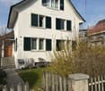 Two-story white house with green shutters, wooden front door, and small garden behind a wooden fence.
