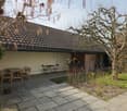 Outdoor patio area with stone tiles, wooden furniture including a table and chairs, small garden plants, and a view of a house with a tiled roof.